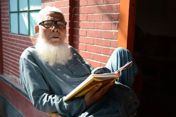 Portrait of bearded man holding book while sitting by brick wall