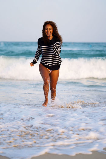 Woman with dark skin and long curly hair having fun at the beach running playing in the waves