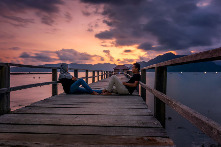 Man and woman sitting on pier over sea against cloudy sky during sunset