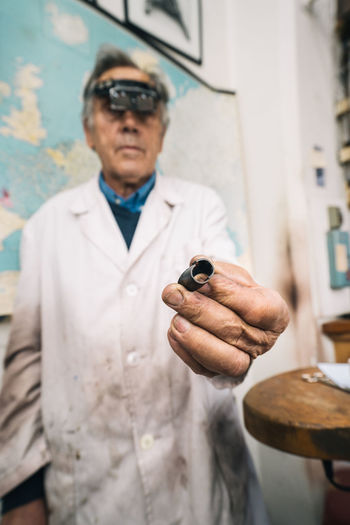 Mature male master in work coat and professional goggles demonstrating metal piece while creating jewelry in workshop