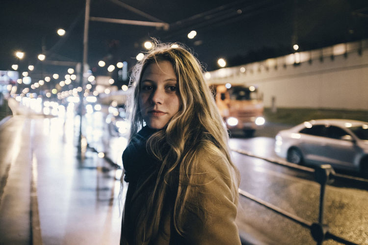 Portrait of beautiful woman standing on illuminated car at night