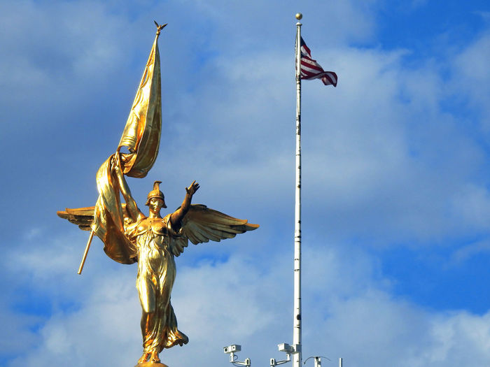 Low angle view of angel statue and flag against blue sky