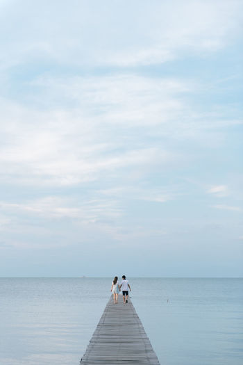 Rear view of couple walking on pier over sea against cloudy sky