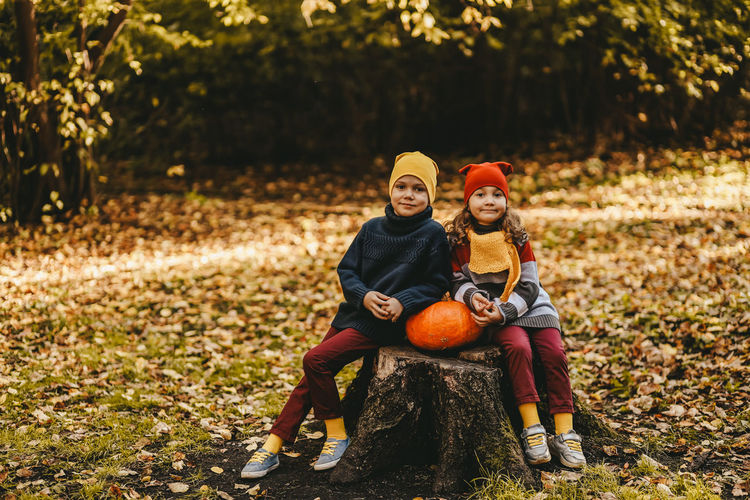 A little girl and a boy children are sitting on a tree stump with a large pumpkin in an autumn park
