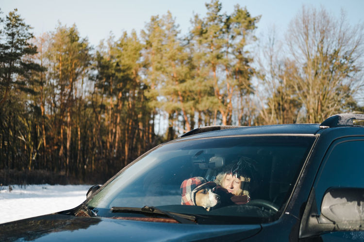 Happy woman with a dog wearing funny winter hats in the car by the forest. 