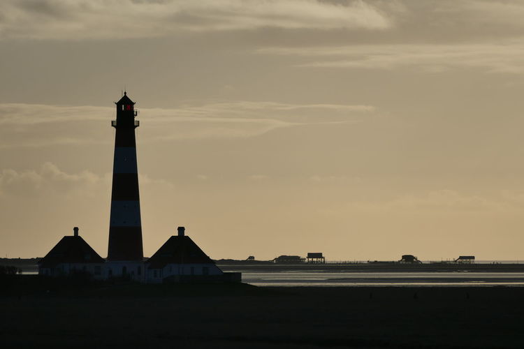 Lighthouse by silhouette building against sky during sunset