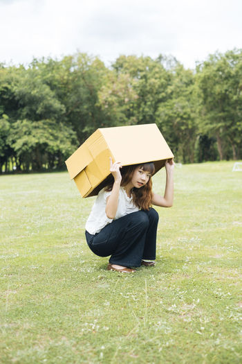 Full length of woman crouching on land while holding cardboard box on grass