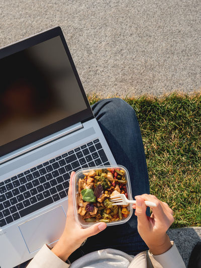 Woman sits on park bench with laptop and  lunch box. bowl with vegetables. healthy nutrition.