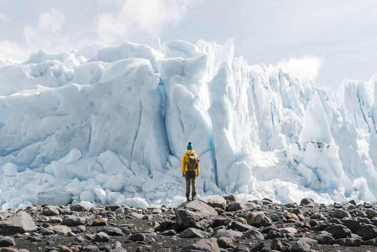 Back view of unrecognizable tourist in outerwear with backpack standing on stony terrain against gigantic blue ice formation while visiting perito moreno glacier in argentina