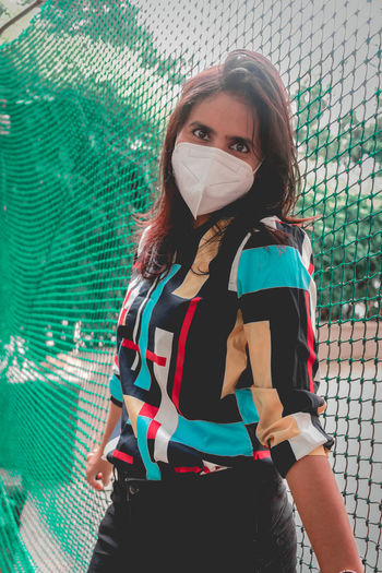 Portrait of woman wearing flu mask standing against net outdoors