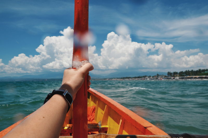 Cropped hand holding pole on boat sailing in sea against cloudy sky