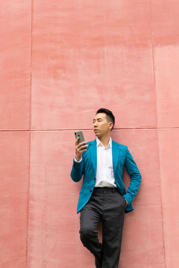 Serious asian male entrepreneur in formal wear browsing on a smartphone looking away while standing on pink background in city