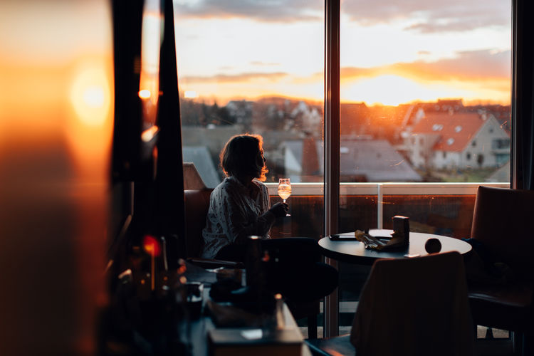 Relaxed woman indoors enjoying a glass of wine and looking to the sunset trough a big window