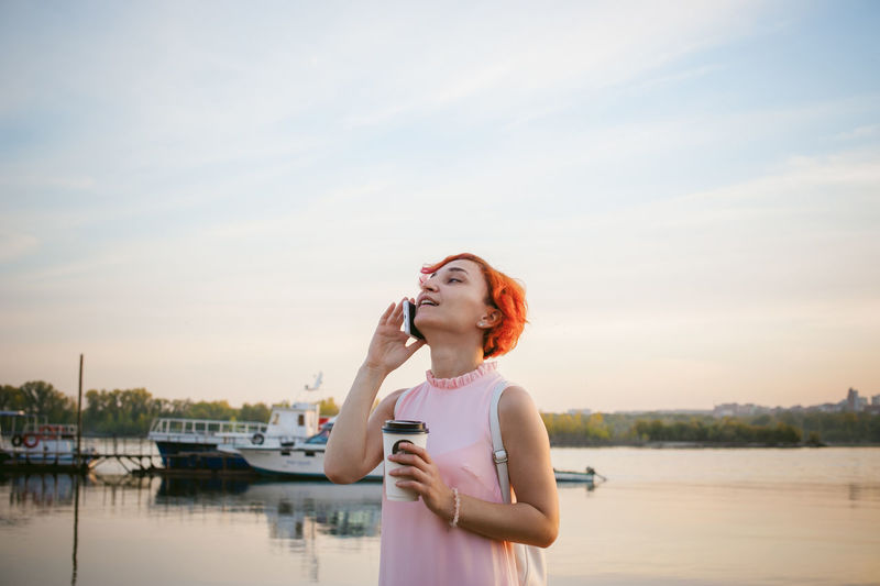 Mid adult woman talking on mobile phone while standing by river against sky