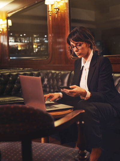 Smart business woman in glasses typing on laptop and checking information in mobile phone comfortably sitting in black leather sofa in cafe