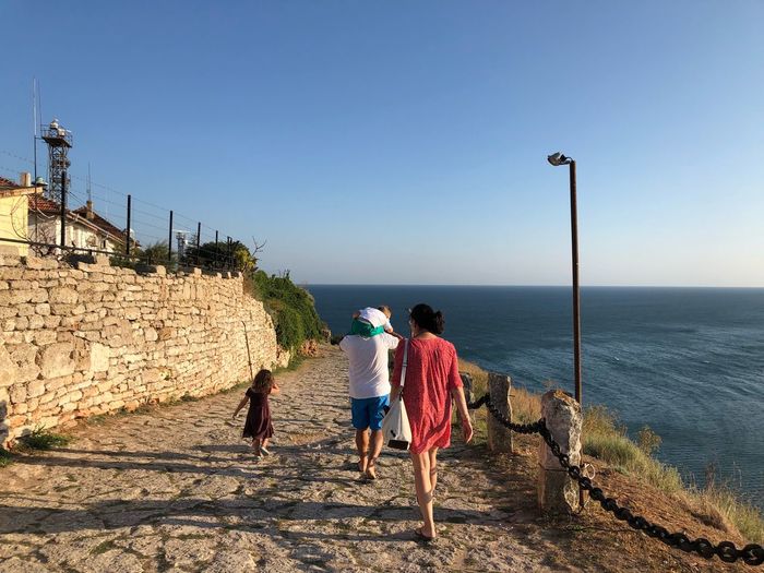 Rear view of family walking on footpath by sea against sky
