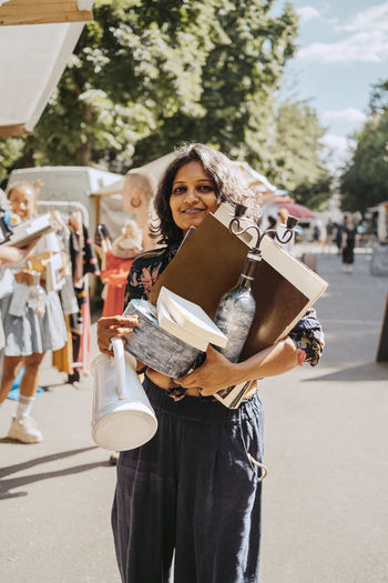Portrait of smiling woman holding various merchandise while standing at flea market