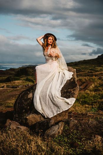 Woman wearing white dress while sitting on rock against cloudy sky