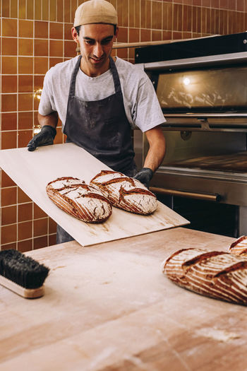 Male baker in apron putting out freshly baked loaves of bread from oven on wooden table in bakehouse