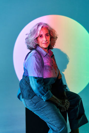 Stylish elderly female in denim clothes with gray hair looking at camera while sitting on block under projection of colorful circle against blue background