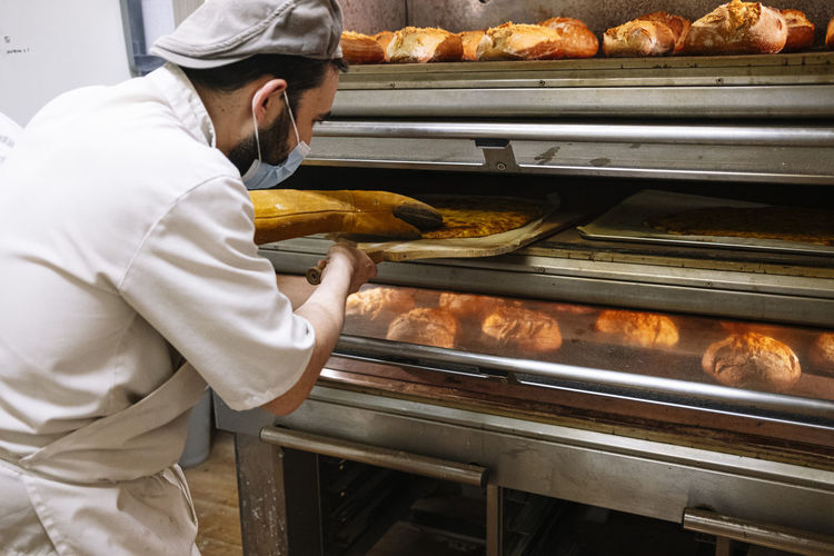 Male baker with pizza peel checking bread in oven at bakery during covid-19