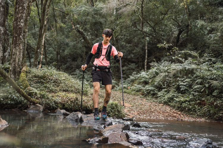 Male traveler with trekking sticks standing near lake in woods