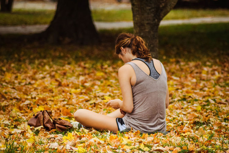 Rear view of woman sitting on fallen dry leaves at park during autumn