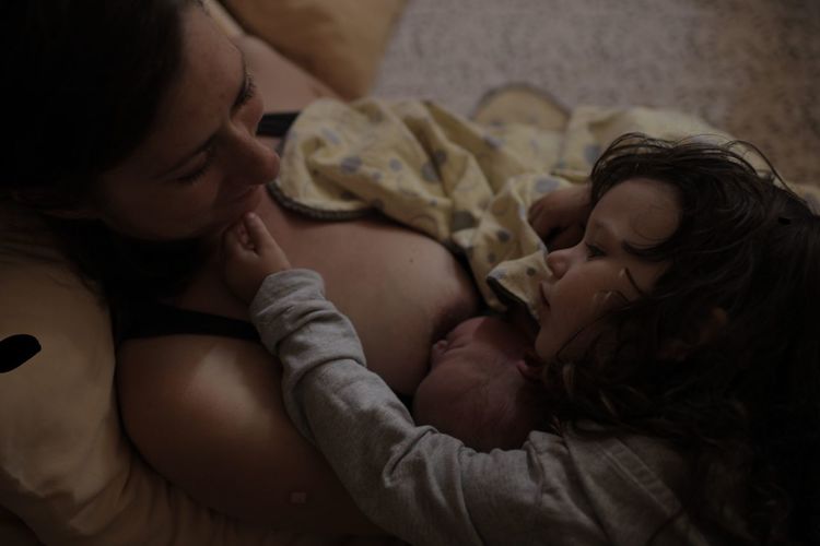 High angle view of girl looking at mother breastfeeding daughter on bed