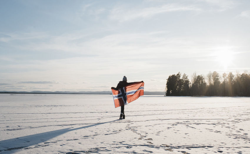 Woman walking across a frozen lake holding a norwegian flag at sunset