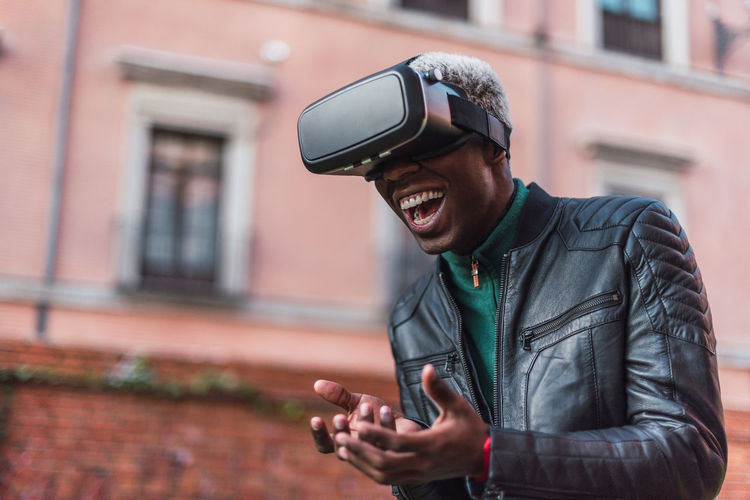 Excited african american male in vr goggles experiencing virtual reality and laughing in city street