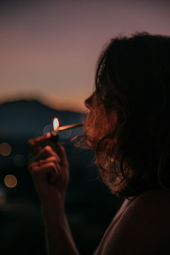 Side view of young faceless lady lighting cigarette with magnificent sunset on blurred background