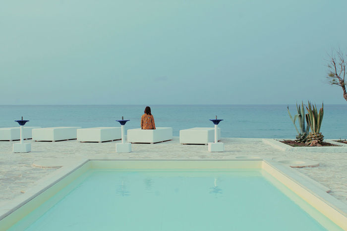 Rear view of woman sitting at poolside by sea against clear sky