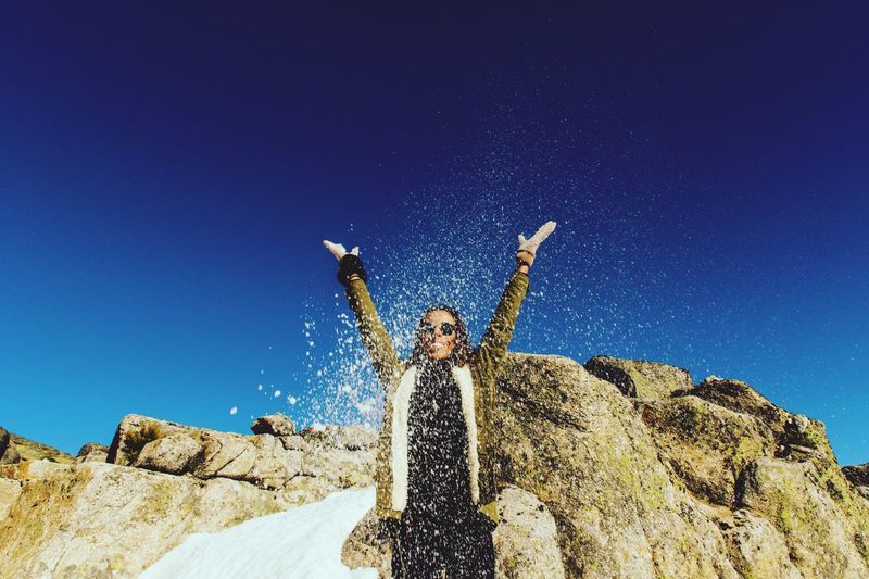 Playful woman throwing sand while standing against rock formations and clear blue sky