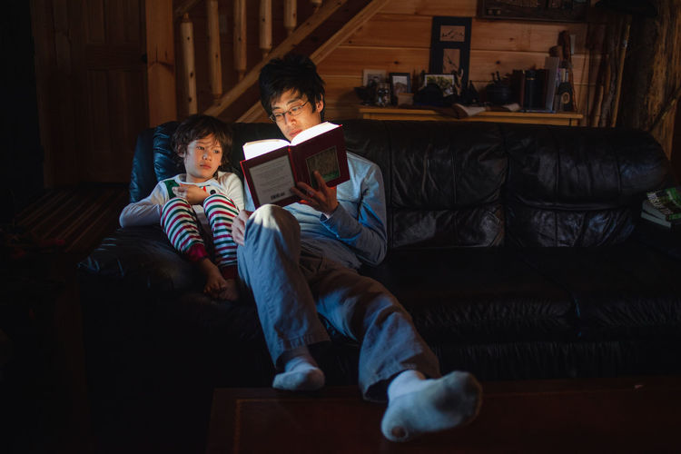 A small boy in pajamas sits with father in early morning reading book