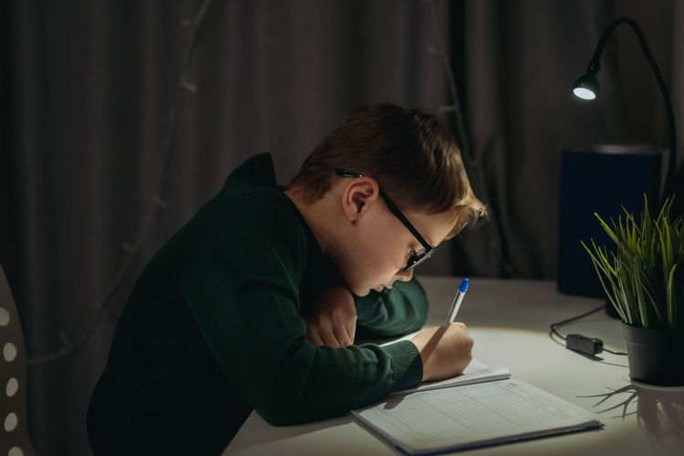 Caucasian boy doing homework at home in dark room by the light of lamp.