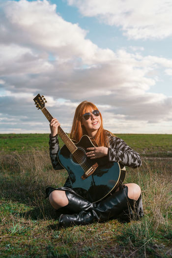 Vertical shot of  caucasian female singer-songwriter with a guitar sitting in the grass of the field