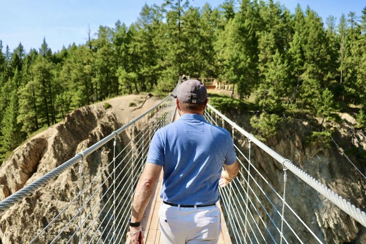 Rear view of man leading the way by railing of suspension bridge against trees