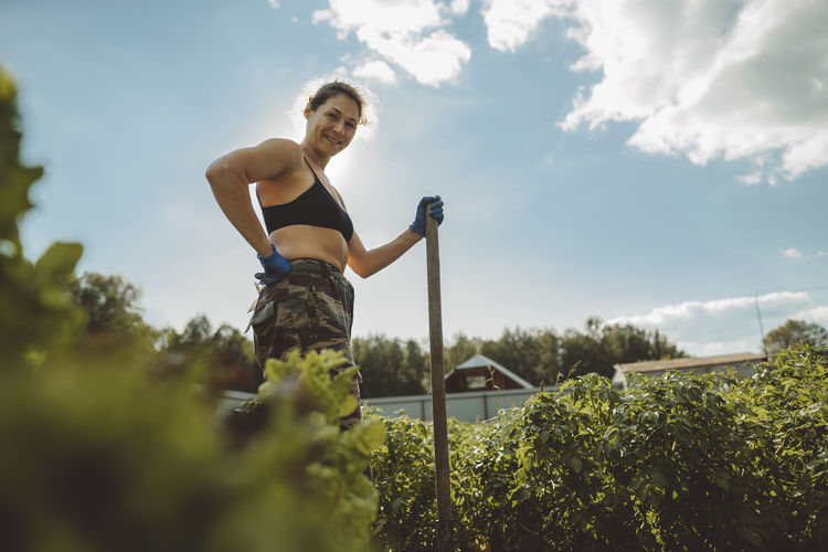 Smiling mature woman holding shovel standing with hand on hip in vegetable garden