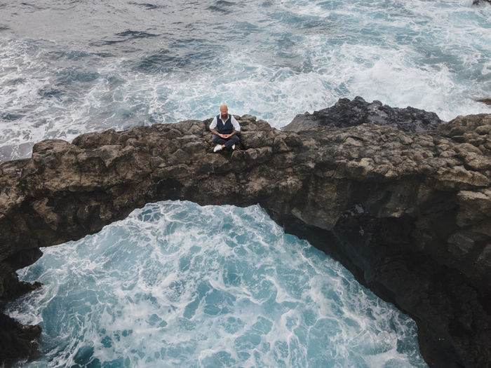 Mature man practicing meditation on rock arch above sea
