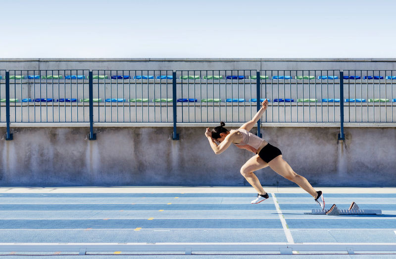 Side view of young strong sportswoman beginning to run fast from starting blocks during track and field workout on stadium