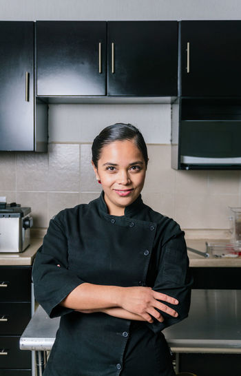 Smiling ethnic female chef in black professional uniform standing at metal table in modern kitchen in restaurant looking at camera