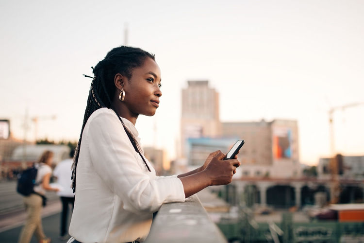 Thoughtful young woman looking away while holding mobile phone on bridge in city