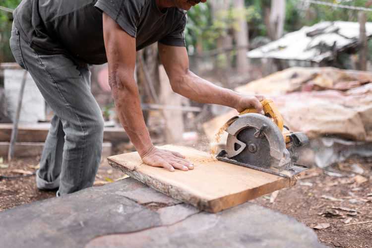 Crop anonymous male worker cutting wooden plank with special grinder on stone surface while working in countryside on summer day