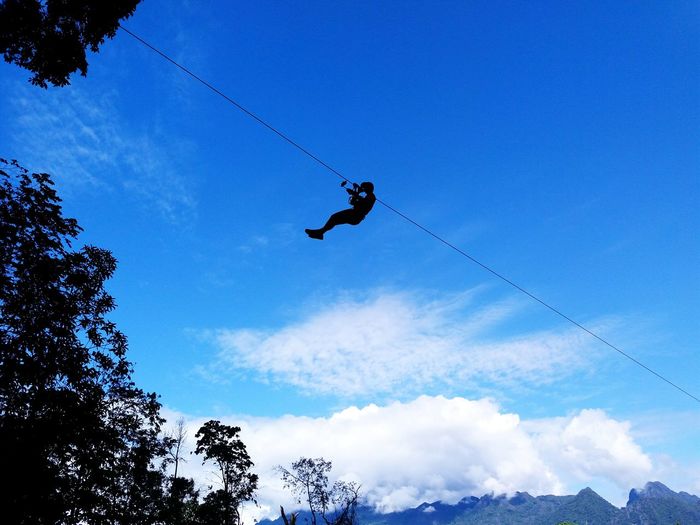 Low angle view of silhouette person zip lining against blue sky