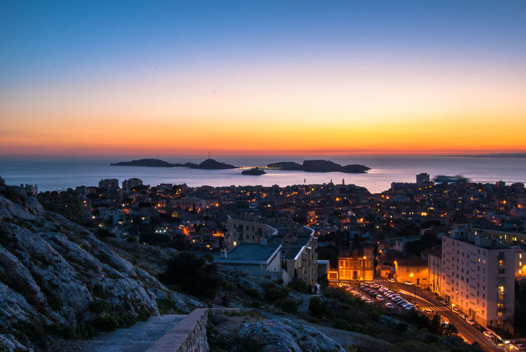 High angle view of illuminated city by sea against sky during sunset