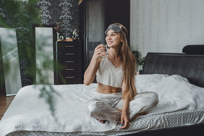 Smiling young woman drinking water while sitting on bed at home