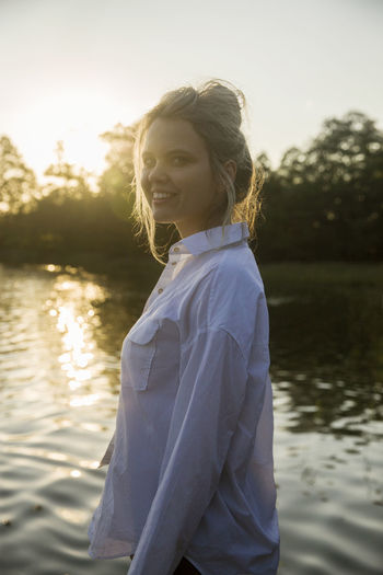 Portrait of smiling young woman standing by lake against sky during sunset