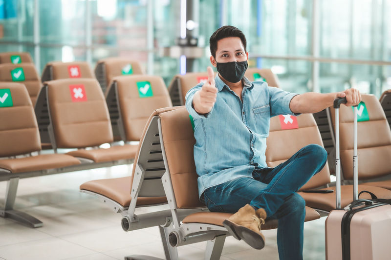 Portrait of mid adult man wearing flu mask gesturing while sitting at airport