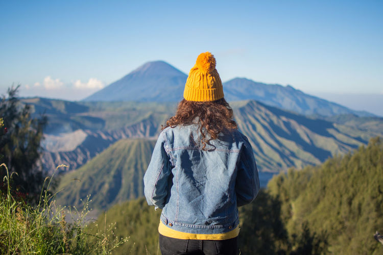 Rear view of woman wearing knit hat and denim jacket while standing on mountain against sky