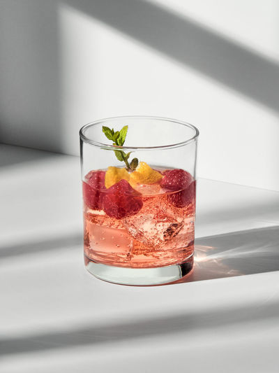 Glass with cocktail with ice cubes and raspberries with mint sprig placed on white surface